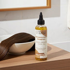 Elama Rosemary Mint Fenugreek Mega Growth Hair Oil bottle on wooden surface with a white bowl holding smooth brown hair
